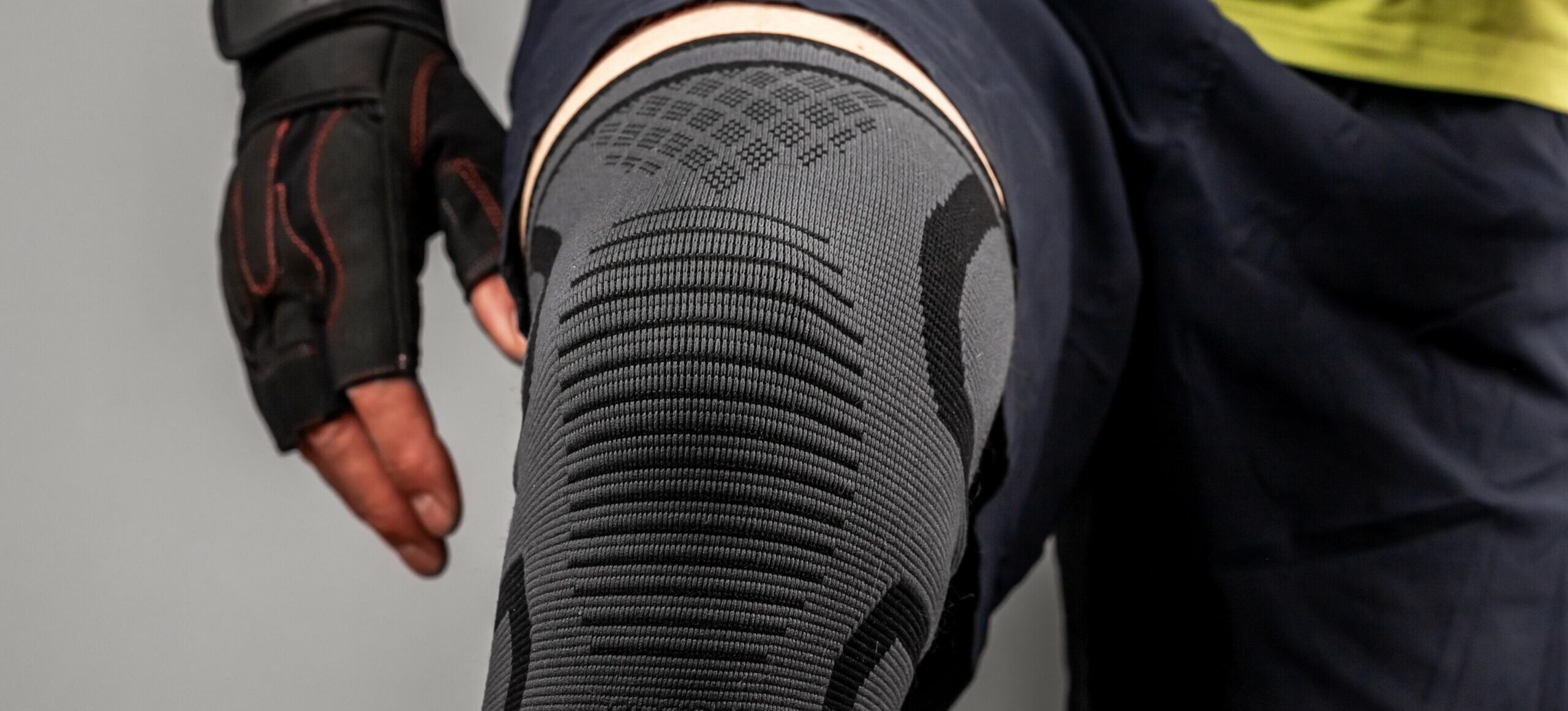 Close-up of a person's knee wearing a grey knitted compression brace. The person is wearing dark athletic shorts and a black fingerless workout glove.