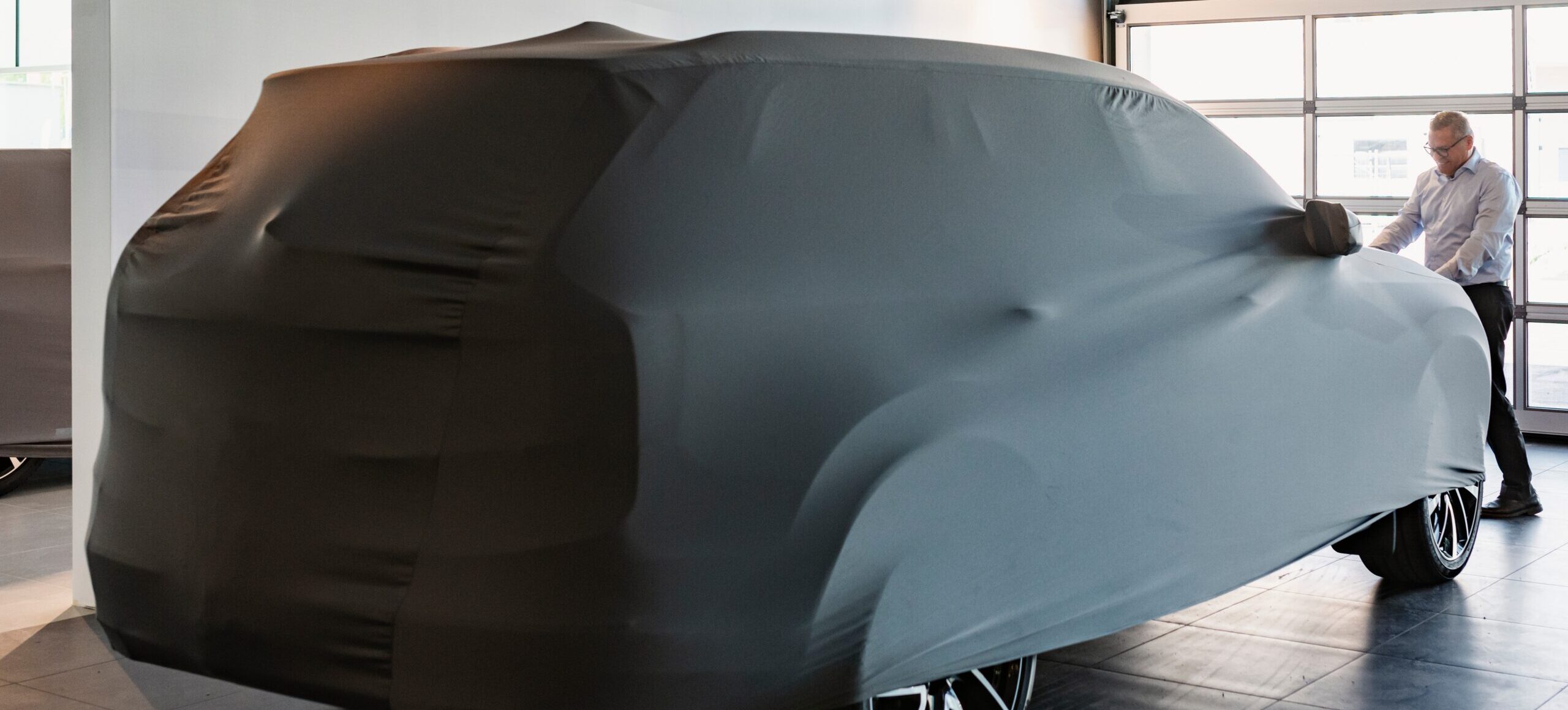 A man in a brightly lit garage or showroom pulling a fitted grey protective cover off of a large vehicle.