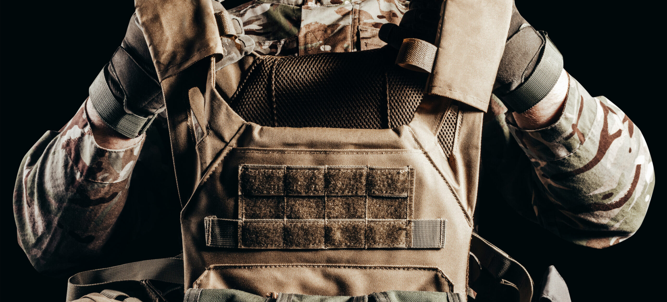 A person in a camouflage uniform adjusting a tan tactical plate carrier vest with modular webbing and pouches, set against a black background.