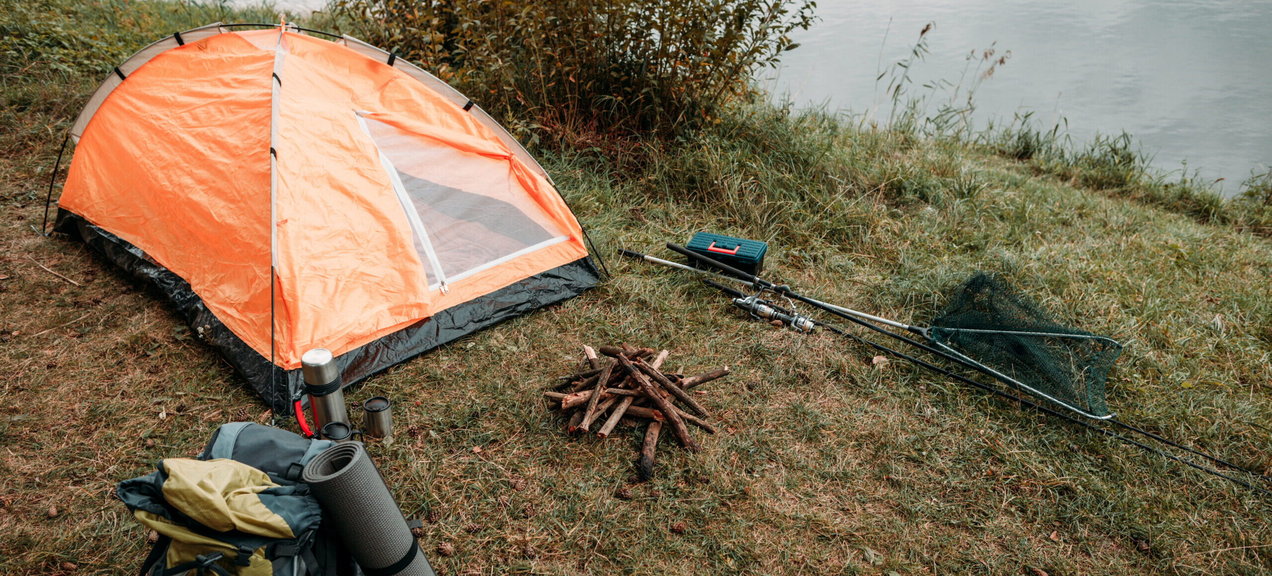 A grassy campsite next to a river featuring a bright orange dome tent, a green backpack with an attached sleeping mat, a small pile of firewood, and fishing equipment.